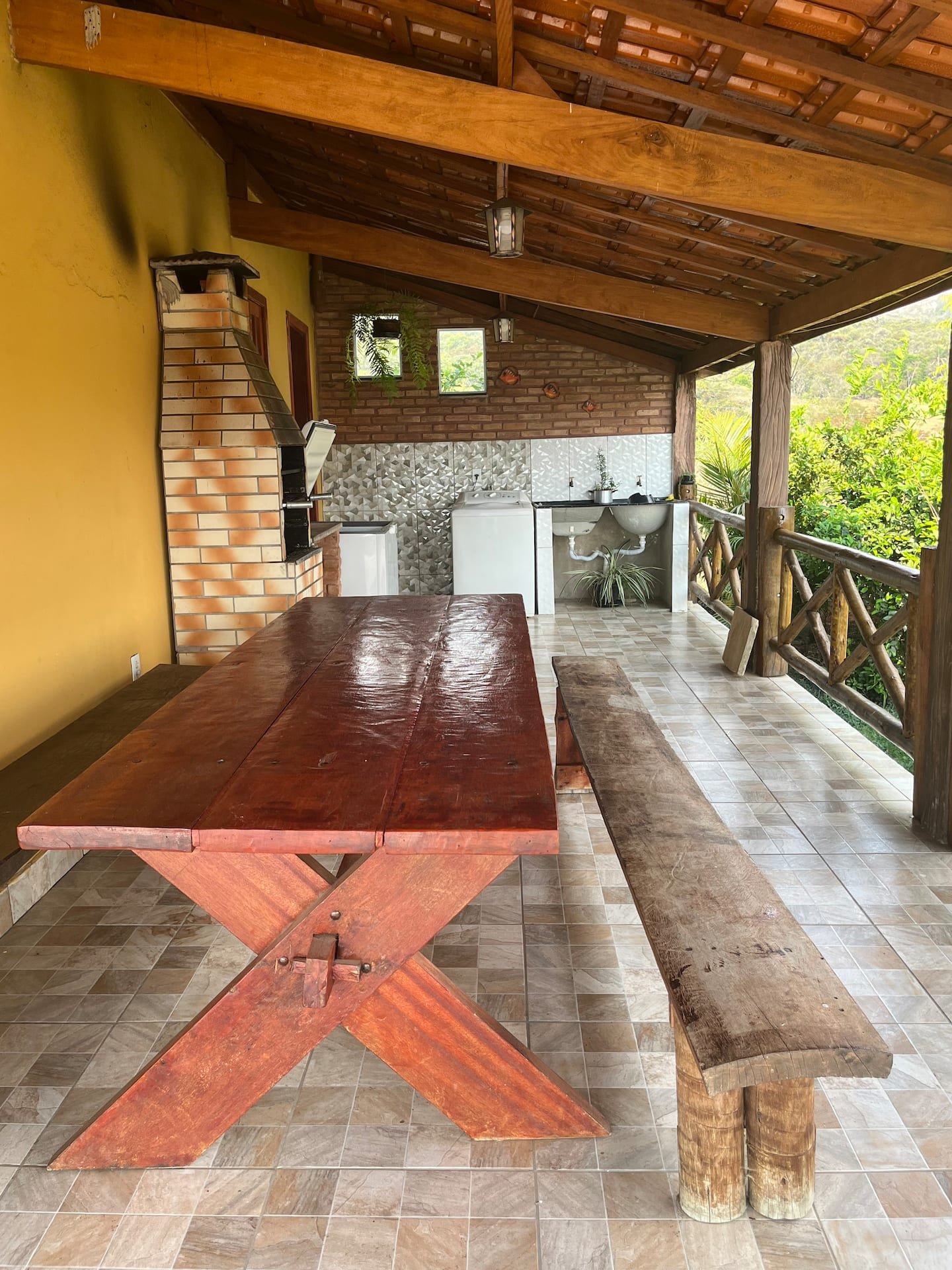 Varanda coberta da casa grande com mesa comprida, bancos e pia ao fundo.
